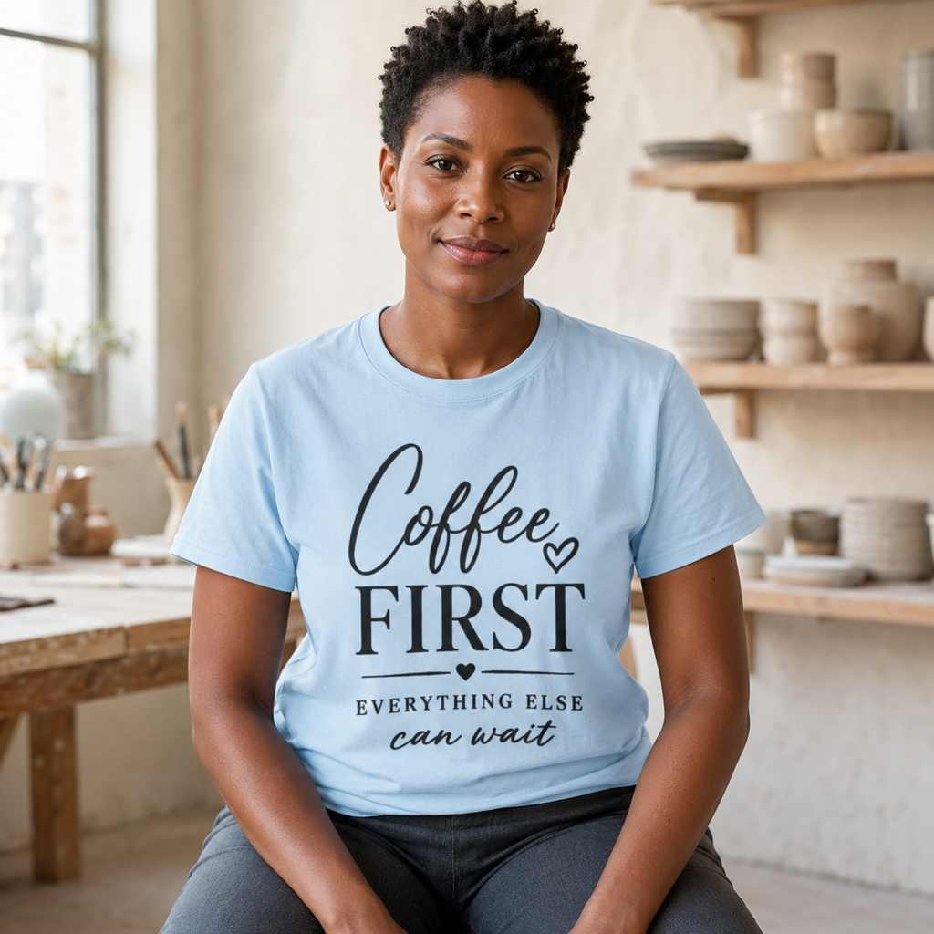 Short-haired Black woman sits in a bright ceramics studio modeling a baby blue T-shirt with clear black typography reading “Coffee First Everything Else Can Wait.” Surrounded by handmade pottery, the image presents a creative maker outfit, pottery lover gift, women’s motivational coffee shirt, relaxed weekend tee, artistic casual clothing, and soft blue caffeine quote top.