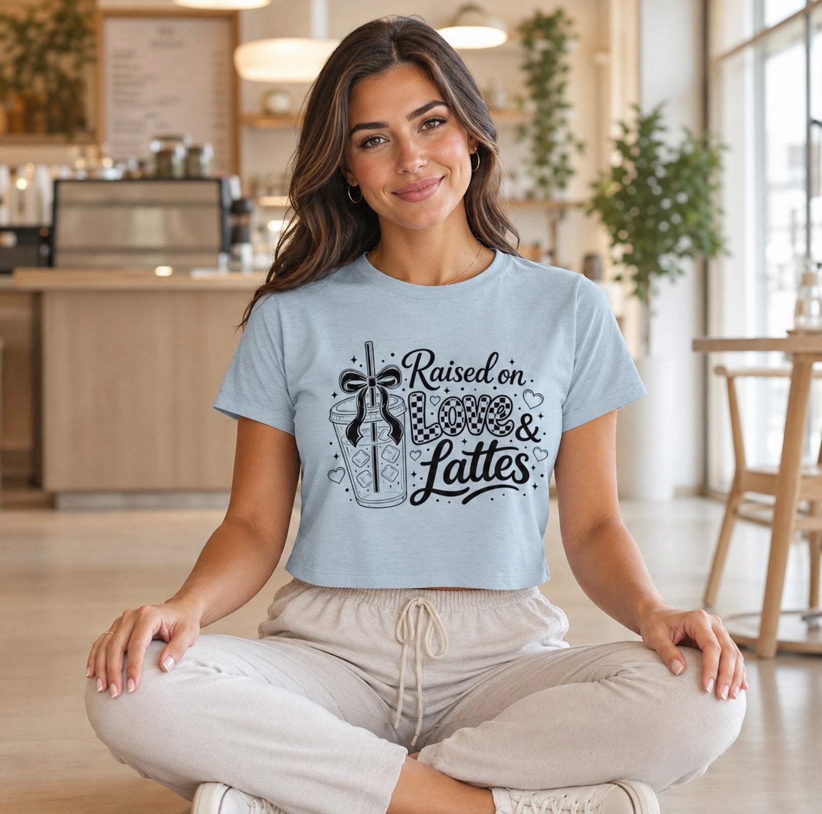 Brunette woman sits cross-legged inside an inviting coffee shop wearing beige pants and a dusty blue “Raised on Love & Lattes” graphic crop shirt. The clear front design showcases flowing script, checkered details and a beverage illustration. Comfortable espresso lover tee for bookstore trips, morning meetups and everyday wear or a creative gift for teachers, nurses and coworkers.