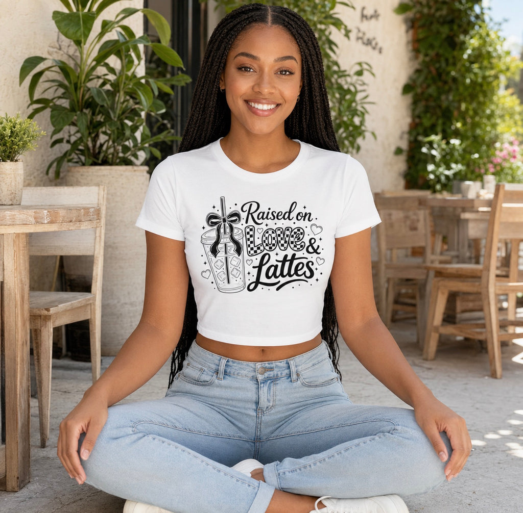 Beautiful Black woman sits cross-legged in a plant-filled coffee shop wearing a fitted white “Raised on Love & Lattes” women’s graphic crop top. The readable black print includes a feminine bow, checkered heart and café beverage artwork. Cute summer shirt for weekend outings, coffee runs and relaxed streetwear or a thoughtful present for a wife, daughter or latte-loving friend.