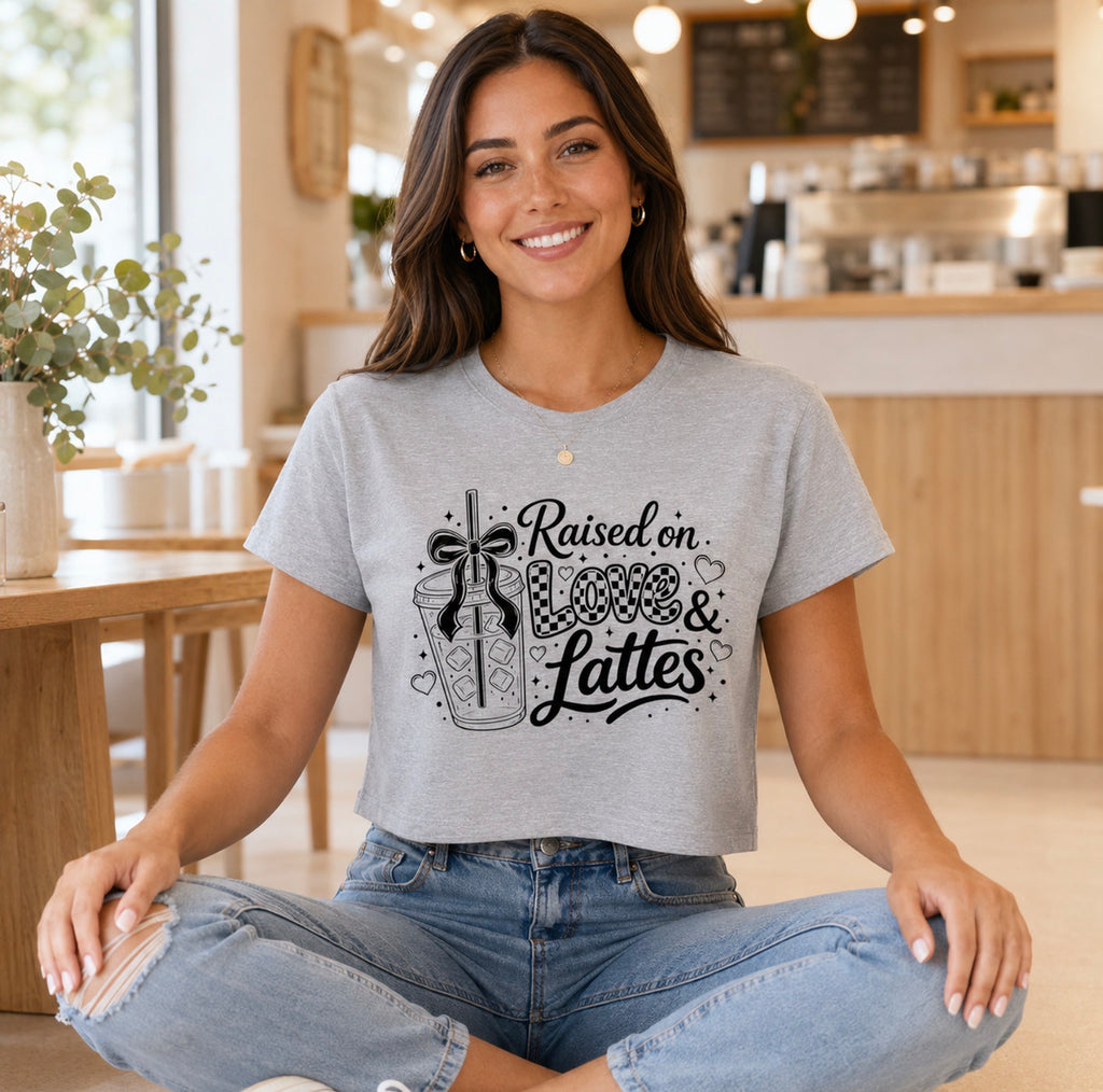 Smiling brunette poses on the floor of a warm café wearing a light gray cropped T-shirt displaying the black “Raised on Love & Lattes” slogan. This modern coffee-themed shirt is made for women who love cappuccinos, cold brew and cute casual fashion. Pair it with denim shorts for an effortless weekend look or choose it as a fun gift for café lovers and caffeine-fueled moms.