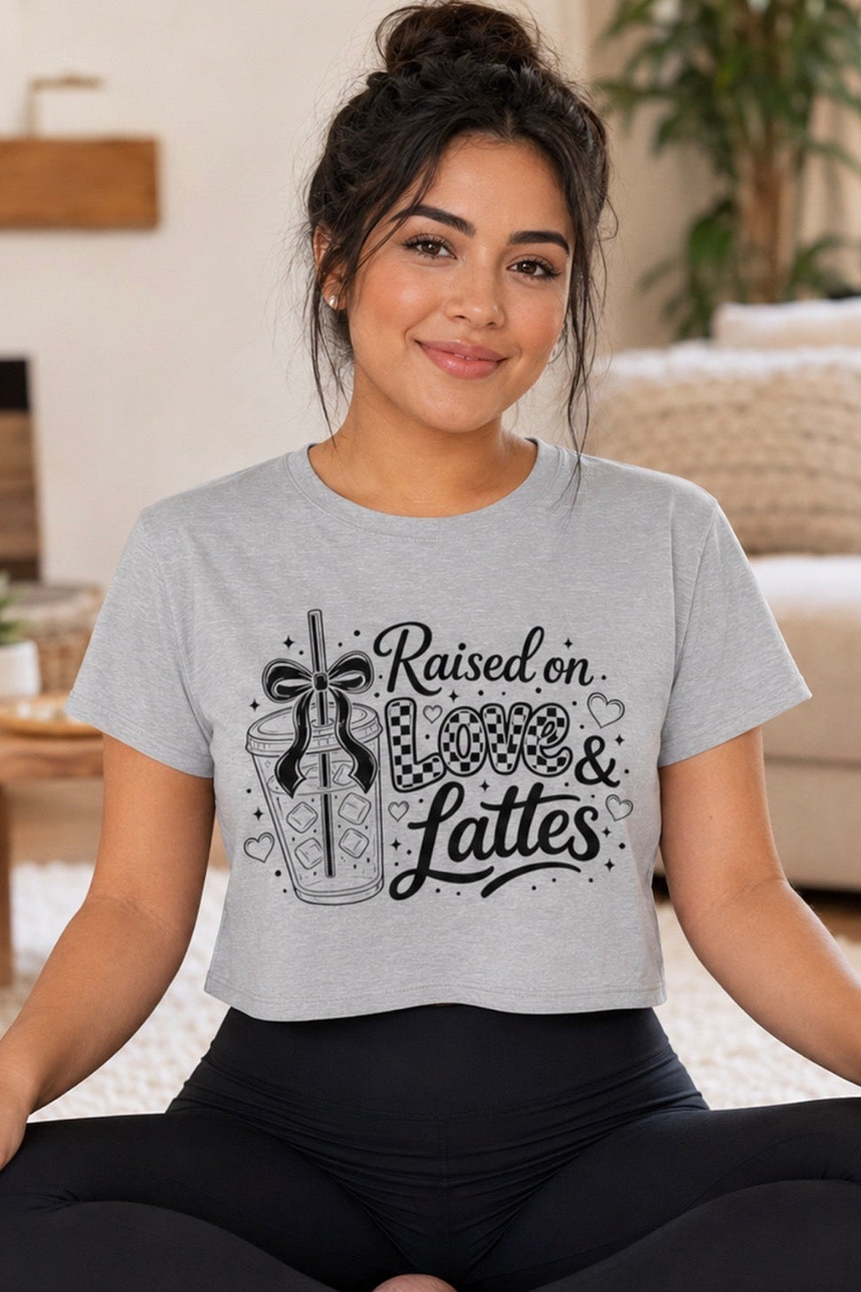 Brunette woman with a messy bun sits cross-legged in a cozy living room wearing a heather gray “Raised on Love & Lattes” cropped graphic tee. This casual women’s coffee shirt features bold black lettering, a bow and drink illustration. Comfortable mom-life top for lounging, running errands or grabbing espresso, plus a cute Mother’s Day, birthday or holiday gift for busy moms.