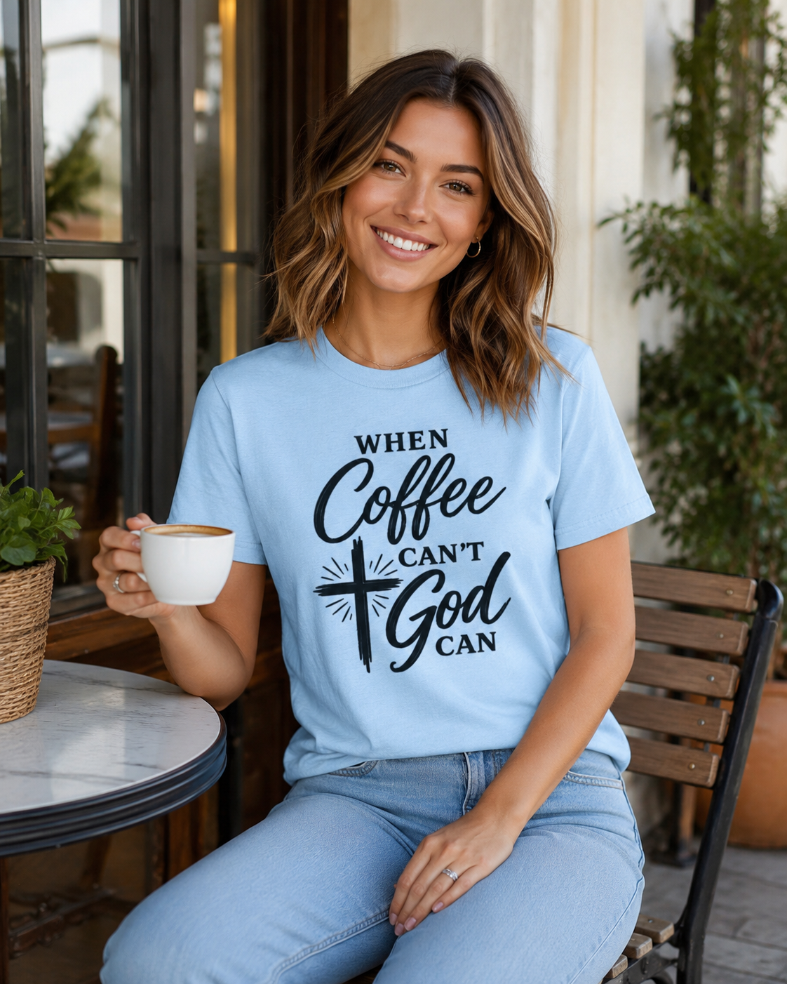 A woman sitting at an outdoor cafe table wearing a light blue graphic t-shirt with the text 'WHEN Coffee CAN'T God CAN' in black lettering, holding a white coffee mug.