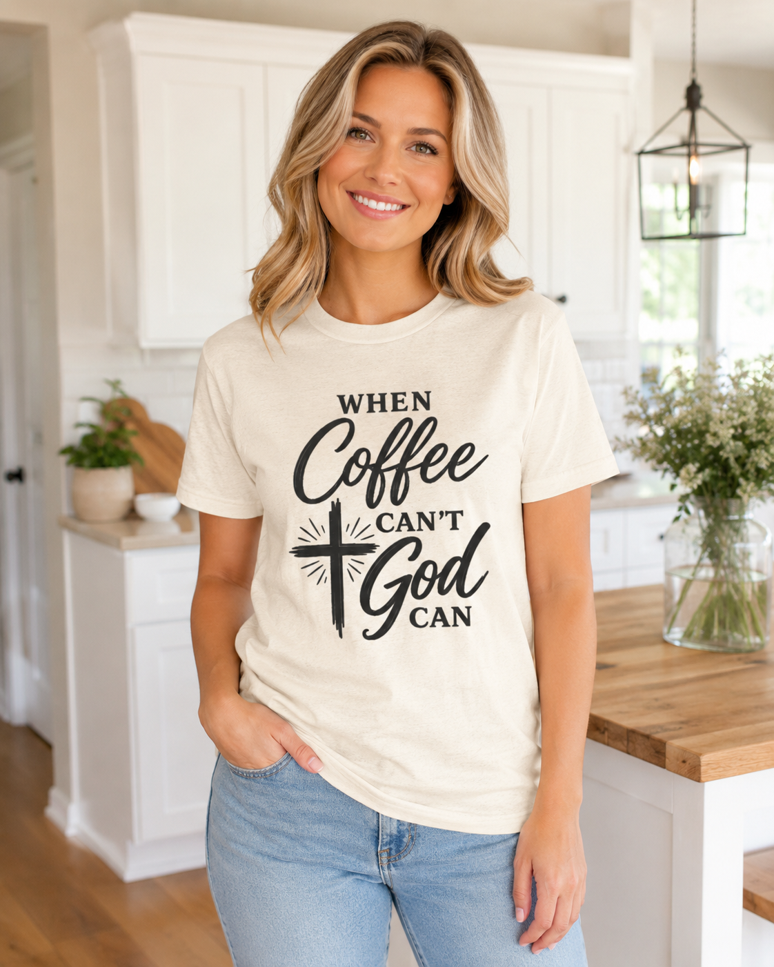 A smiling blonde woman wearing a cream-colored graphic t-shirt with the text 'WHEN Coffee CAN'T God CAN' in black, paired with light blue jeans, standing in a bright white kitchen.