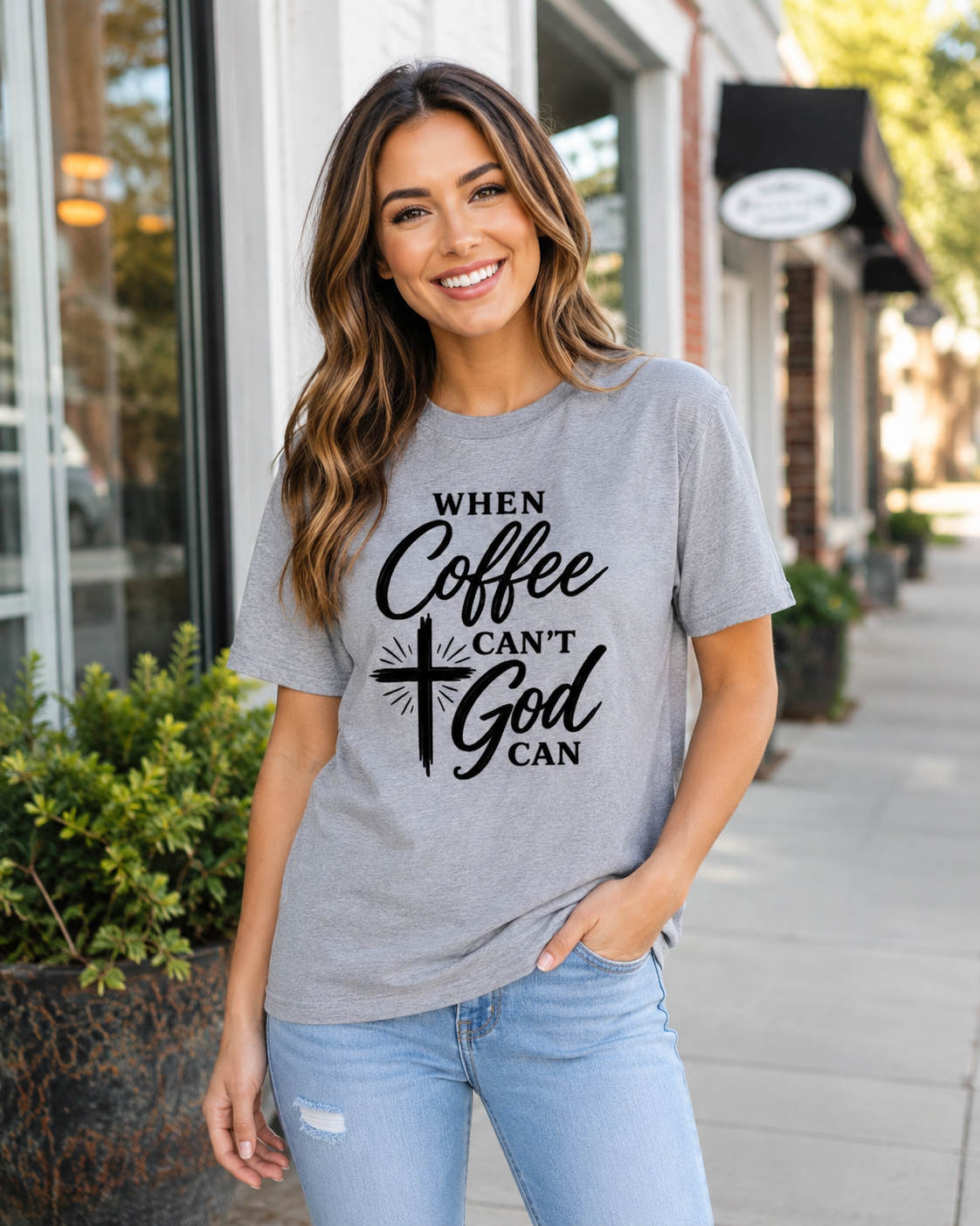 A woman wearing a heather gray graphic t-shirt with the text 'WHEN Coffee CAN'T God CAN' in black script and cross design, paired with light blue jeans, standing on a city sidewalk.