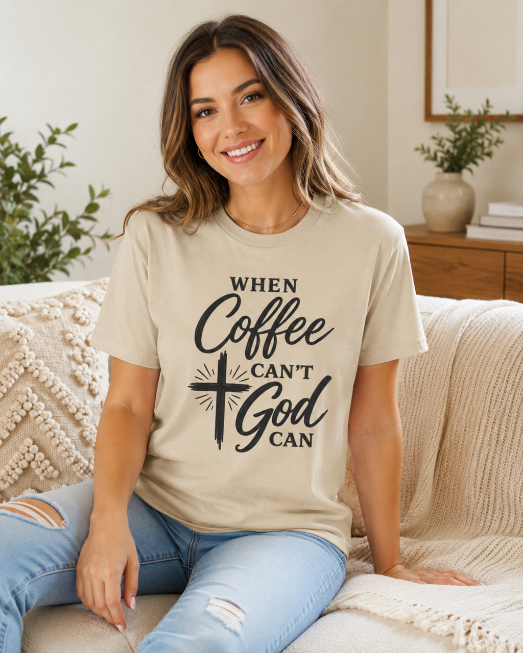 A woman sitting on a cozy beige couch wearing a beige graphic t-shirt with the text 'WHEN Coffee CAN'T God CAN' in black script and block lettering, paired with light blue ripped jeans.