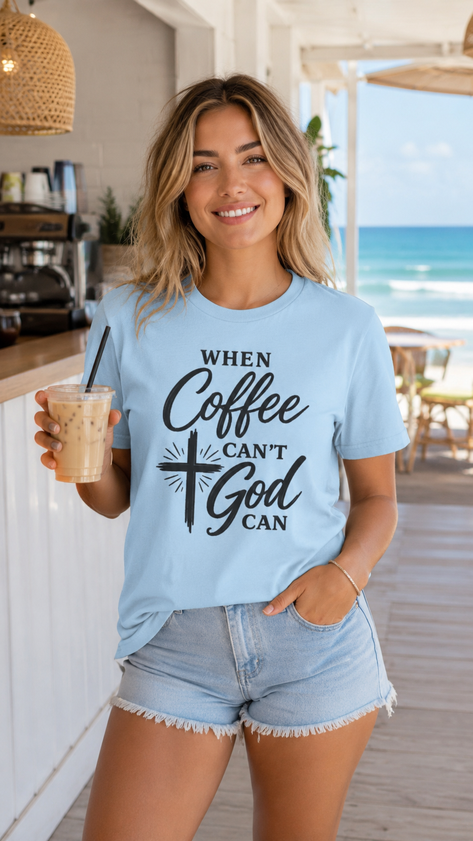 A woman wearing a light blue graphic t-shirt with the text 'WHEN Coffee CAN'T God CAN' in black, holding an iced coffee in a bright, modern cafe setting with a beach view in the background.