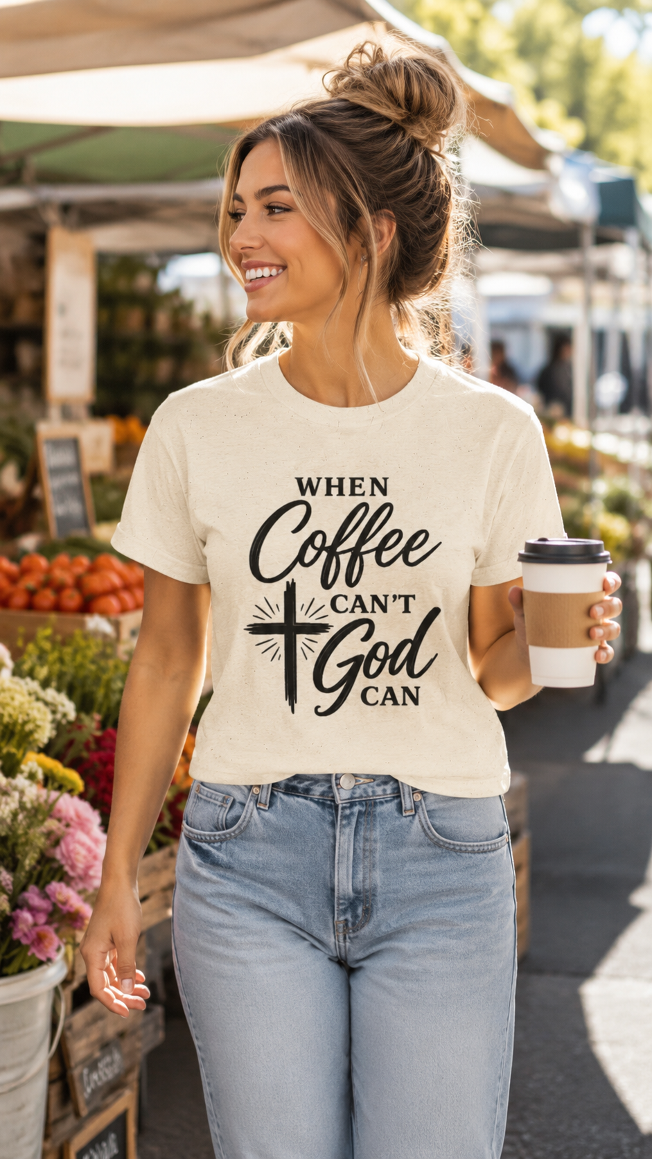A woman wearing a cream-colored graphic t-shirt with the text 'WHEN Coffee CAN'T God CAN' in black script and a cross graphic, paired with light-wash blue jeans, holding a coffee cup in an outdoor market setting.