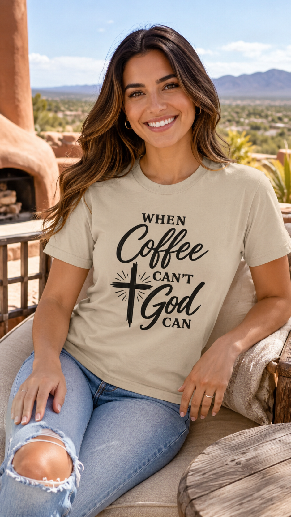 A woman sitting outdoors on a patio wearing a beige graphic t-shirt with the text 'WHEN Coffee CAN'T God CAN' in black script and print, paired with distressed blue jeans.