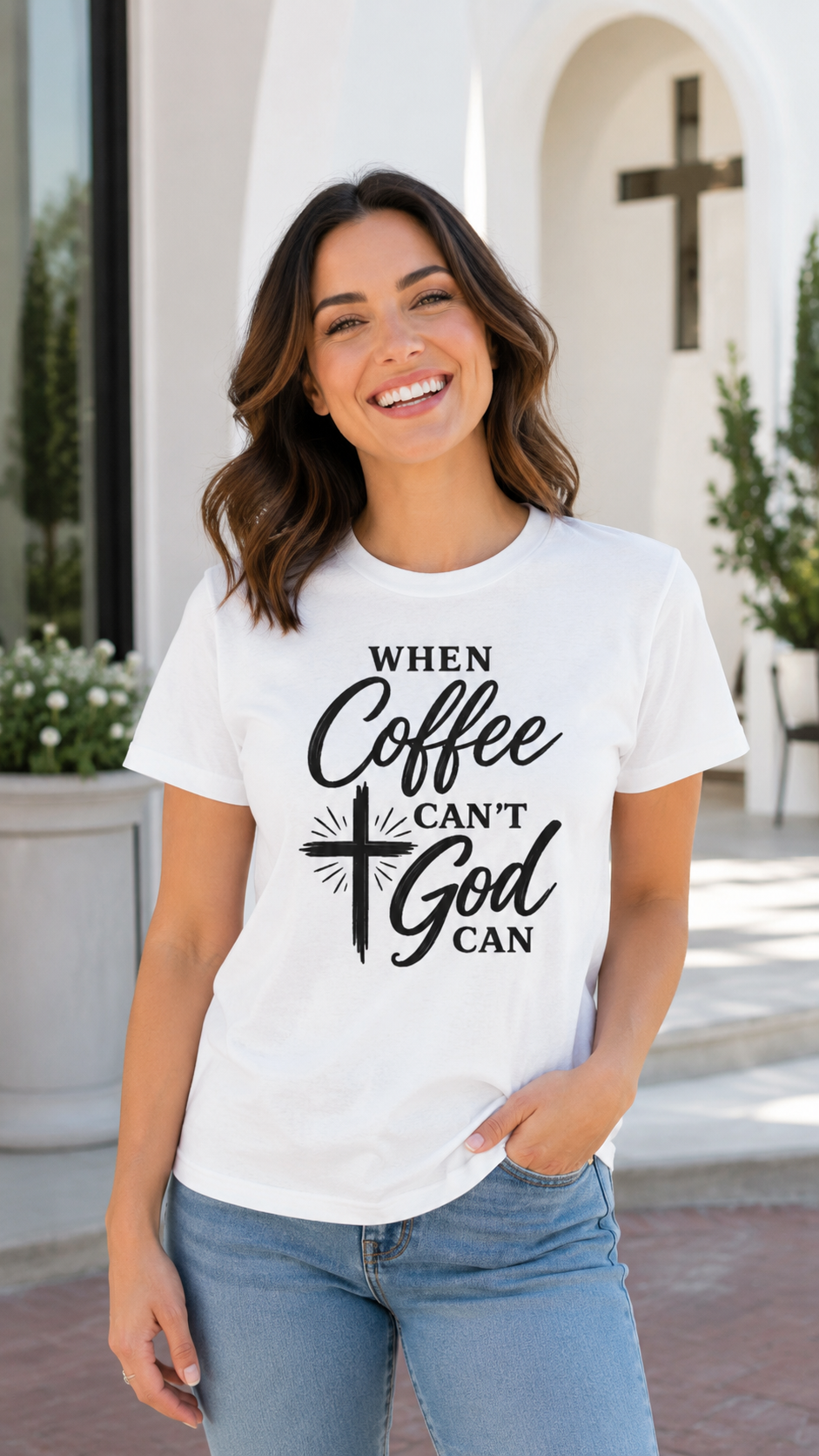 A smiling woman wearing a white graphic t-shirt with the text 'WHEN Coffee CAN'T God CAN' in black script and cross design, paired with blue jeans, standing outdoors in front of a white building with a cross.
