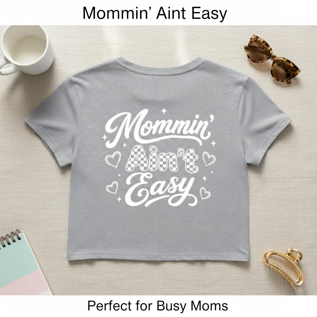 Gray women’s Mommin Ain’t Easy crop top with white mom graphic shown with coffee cup and sunglasses props.