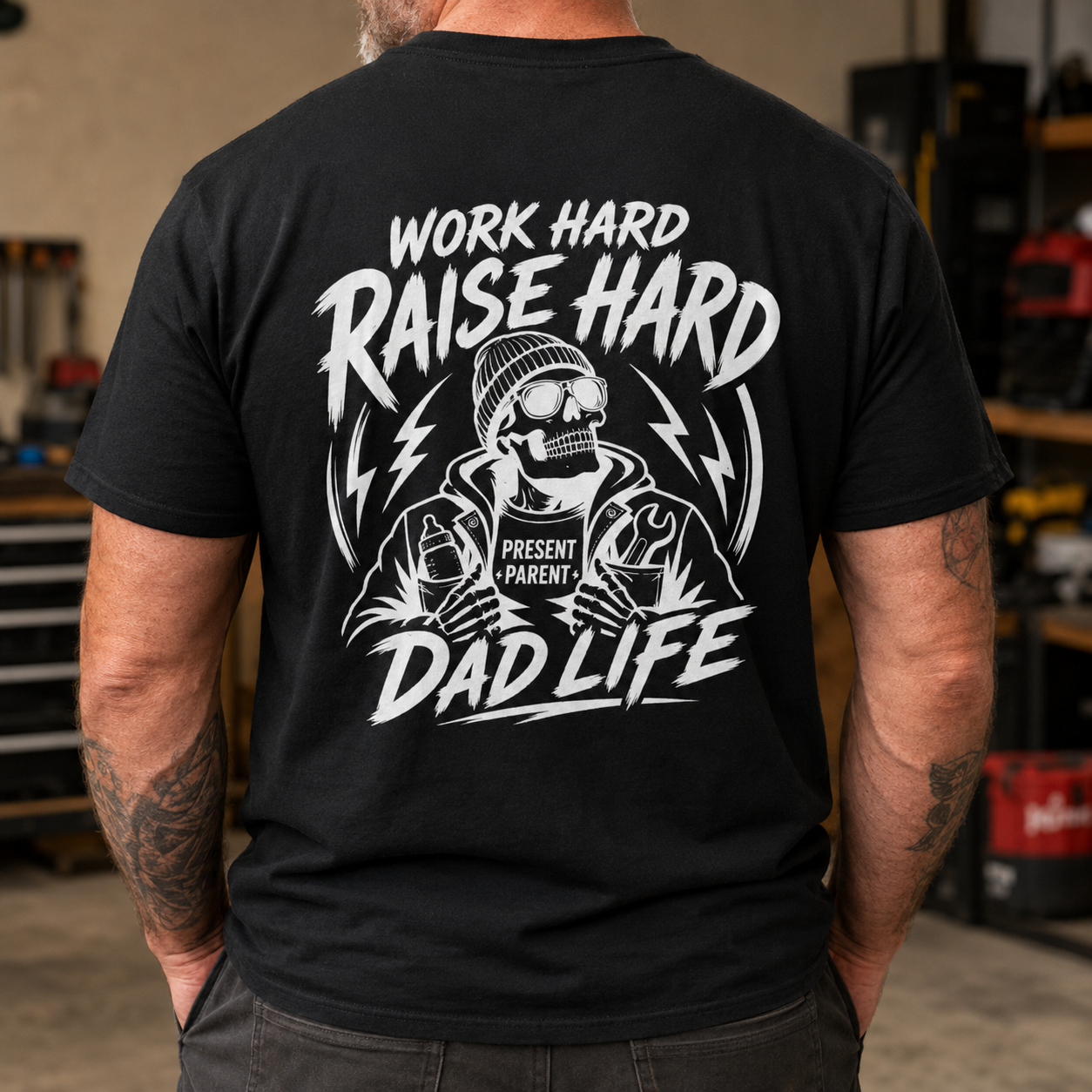 Black men’s graphic T-shirt modeled from the back inside a rugged workshop, featuring a bold white skeleton design with the words “Work Hard Raise Hard Dad Life” and PresentParent detail. Blue collar dad shirt, working father apparel, construction dad gift, masculine skull tee, Father’s Day present and TikTok Shop workwear for hardworking dads.