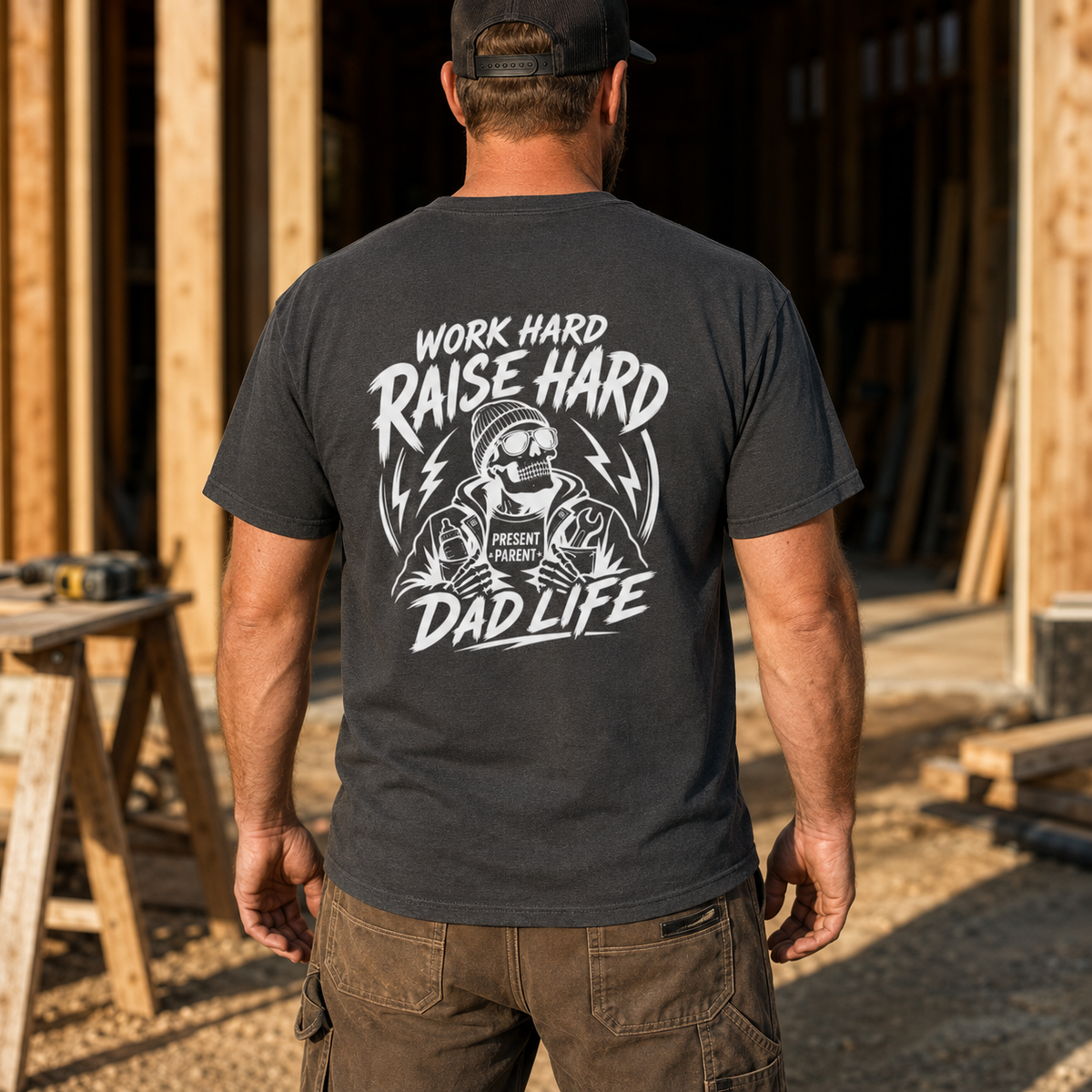 Charcoal back-print tee modeled by a man walking through a lumber work area, featuring the large white “Work Hard Raise Hard Dad Life” skeleton design. Contractor uniform style, framing carpenter shirt, home builder apparel, gritty jobsite fashion, gift for foreman dad, men’s casual work top and meaningful clothing honoring fathers building careers, homes and futures.