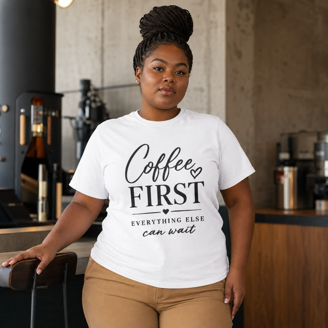 Plus-size Black woman with a braided high bun models a crisp white T-shirt inside a contemporary coffee roastery. The bold black front design says “Coffee First Everything Else Can Wait” above camel pants. Ideal search details include plus size coffee shirt, barista gift for women, espresso lover apparel, café-inspired outfit, white statement tee, and coffee shop worker gift.