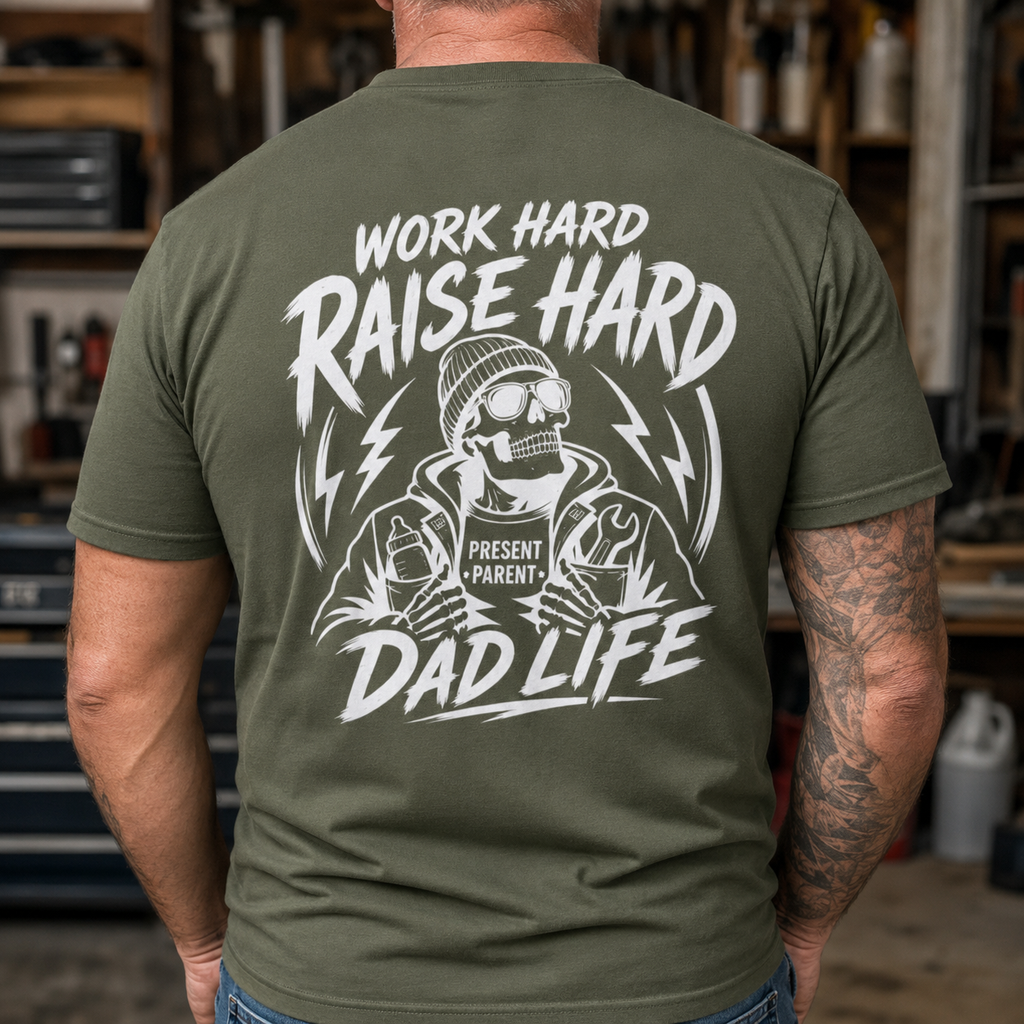 A man viewed from behind wearing an olive green t-shirt with a large white graphic on the back that reads 'WORK HARD RAISE HARD DAD LIFE' and features a skeleton dad illustration. The background is a blurred workshop environment.