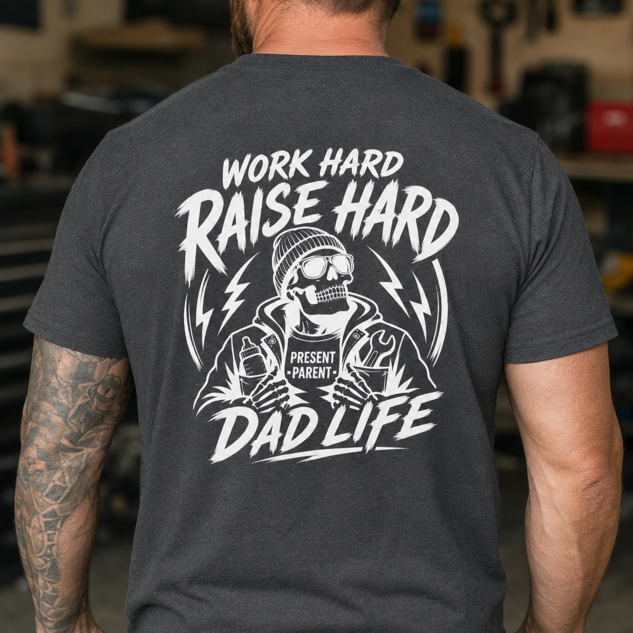 Charcoal gray graphic shirt worn by a tattooed man in a garage setting, showcasing the oversized white skeleton and “Work Hard Raise Hard Dad Life” back artwork. Mechanic dad tee, tattoo style menswear, garage work shirt, cool gift for fathers, skull graphic clothing, hardworking husband present and edgy TikTok fashion for proud blue collar dads.
