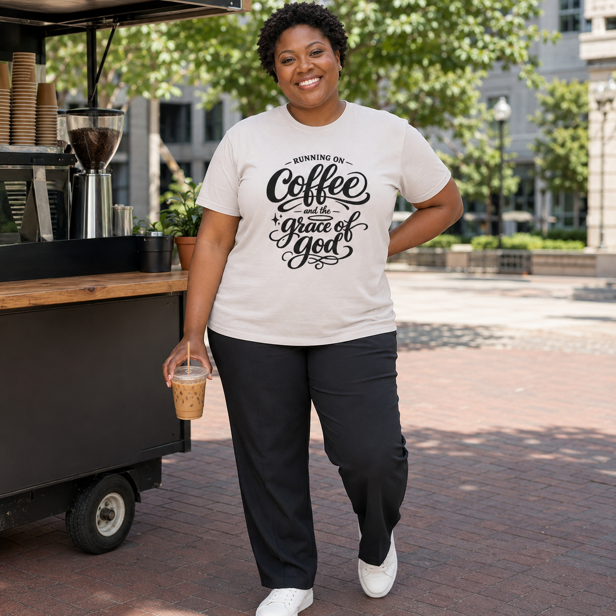 A woman stands outdoors in front of a coffee cart, wearing a cream graphic T-shirt with black script text reading 'Running on Coffee and the grace of God' and dark pants.