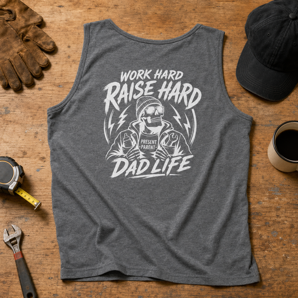 A heather dark gray sleeveless tank top laid flat on a wooden surface, featuring a large white graphic with the text 'WORK HARD RAISE HARD DAD LIFE' and an illustrated dad figure. Surrounding props include gloves, a cap, a coffee cup, a tape measure, and a wrench.