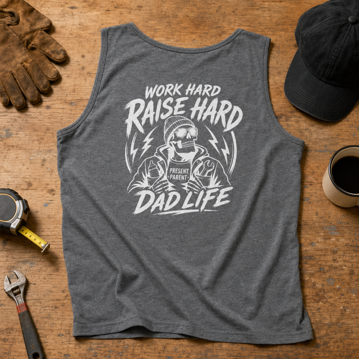 A heather dark gray sleeveless tank top laid flat on a wooden surface, featuring a large white graphic with the text 'WORK HARD RAISE HARD DAD LIFE' and an illustrated dad figure. Surrounding props include gloves, a cap, a coffee cup, a tape measure, and a wrench.