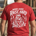 A man viewed from behind wearing a red t-shirt with a large white graphic print that reads 'WORK HARD RAISE HARD DAD LIFE' and features a skeleton dad holding a coffee mug that says 'PRESENT PARENT'.