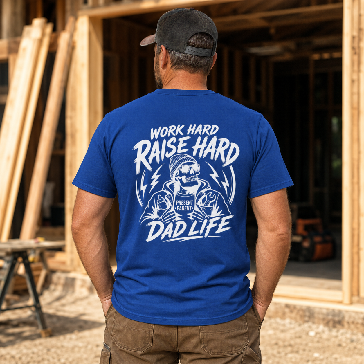 Royal-blue men’s crewneck shirt photographed from the rear near stacked lumber, featuring a crisp white skeleton graphic and “Work Hard Raise Hard Dad Life” message. Carpenter dad gift, blue workwear T-shirt, woodworking father apparel, laborer outfit, dad birthday idea, shop clothing and statement tee celebrating men who grind all day and show up for their children.
