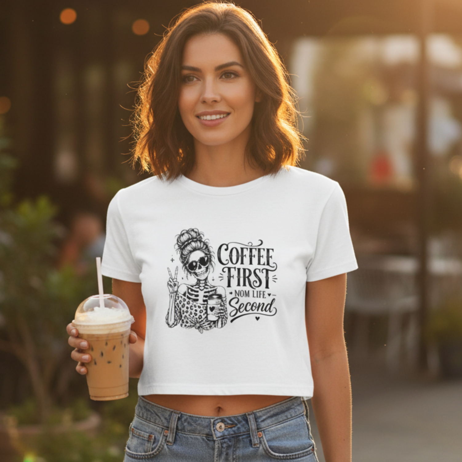 Model wearing a white cropped coffee graphic t-shirt with Coffee First Mom Life Second skeleton print while holding an iced coffee outdoors. Cute women's summer crop top, coffee shop outfit, aesthetic graphic tee, trendy mom clothing, casual weekend fashion, gift for coffee drinkers, stylish everyday apparel.