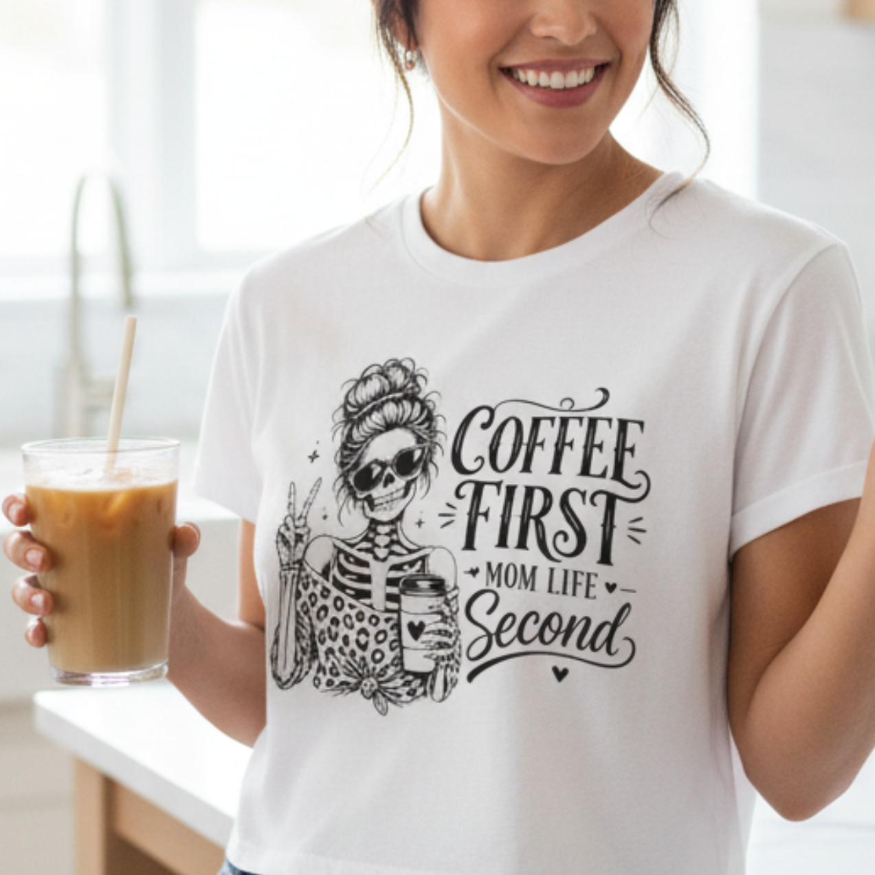 Woman standing in a bright kitchen wearing a white Coffee First Mom Life Second cropped graphic t-shirt with skeleton coffee artwork. Women's casual coffee lover shirt, everyday crop top, trendy mom apparel, boutique graphic tee, soft comfortable fashion, coffee gift idea, modern lifestyle clothing.