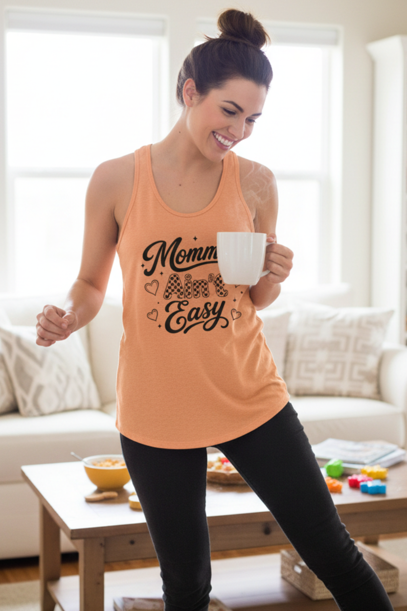 Woman wearing a light orange Mommin’ Ain’t Easy racerback tank top at home, cute coffee lover mom shirt and casual Mother’s Day gift for everyday mom life.