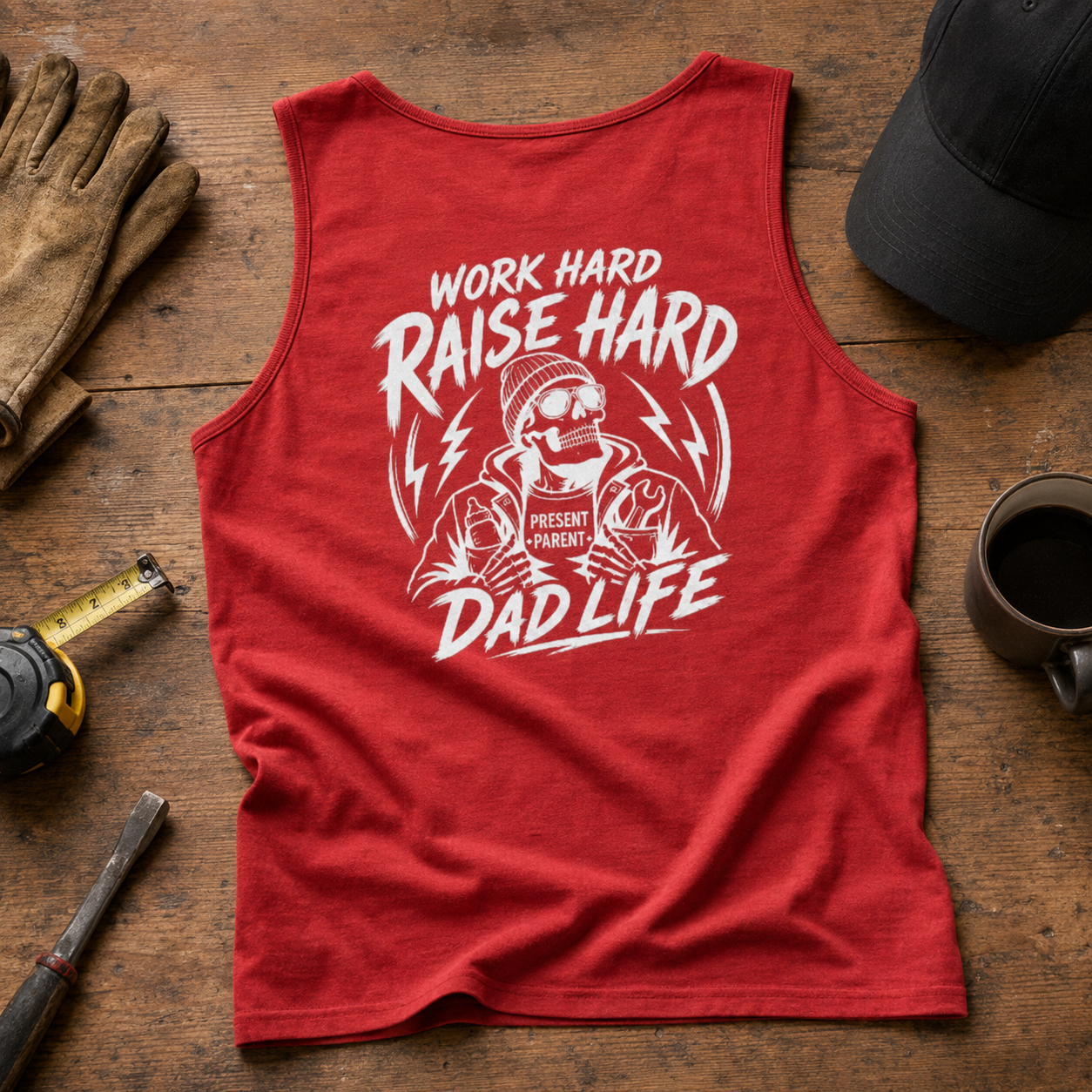 A red sleeveless tank top laid flat on a rustic wooden surface, featuring a large white graphic of a construction worker and the text 'WORK HARD RAISE HARD DAD LIFE.' Surrounding props include work gloves, a tape measure, a hammer, a coffee mug, and a black cap.
