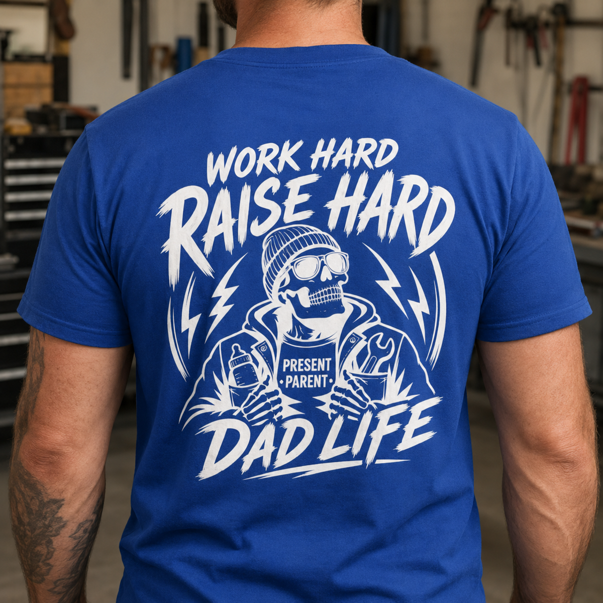 A man viewed from behind wearing a royal blue t-shirt with a large white graphic print on the back. The graphic features a skeleton wearing sunglasses and a beanie, holding coffee cups, with the text 'WORK HARD RAISE HARD DAD LIFE' and 'PRESENT PARENT+'.